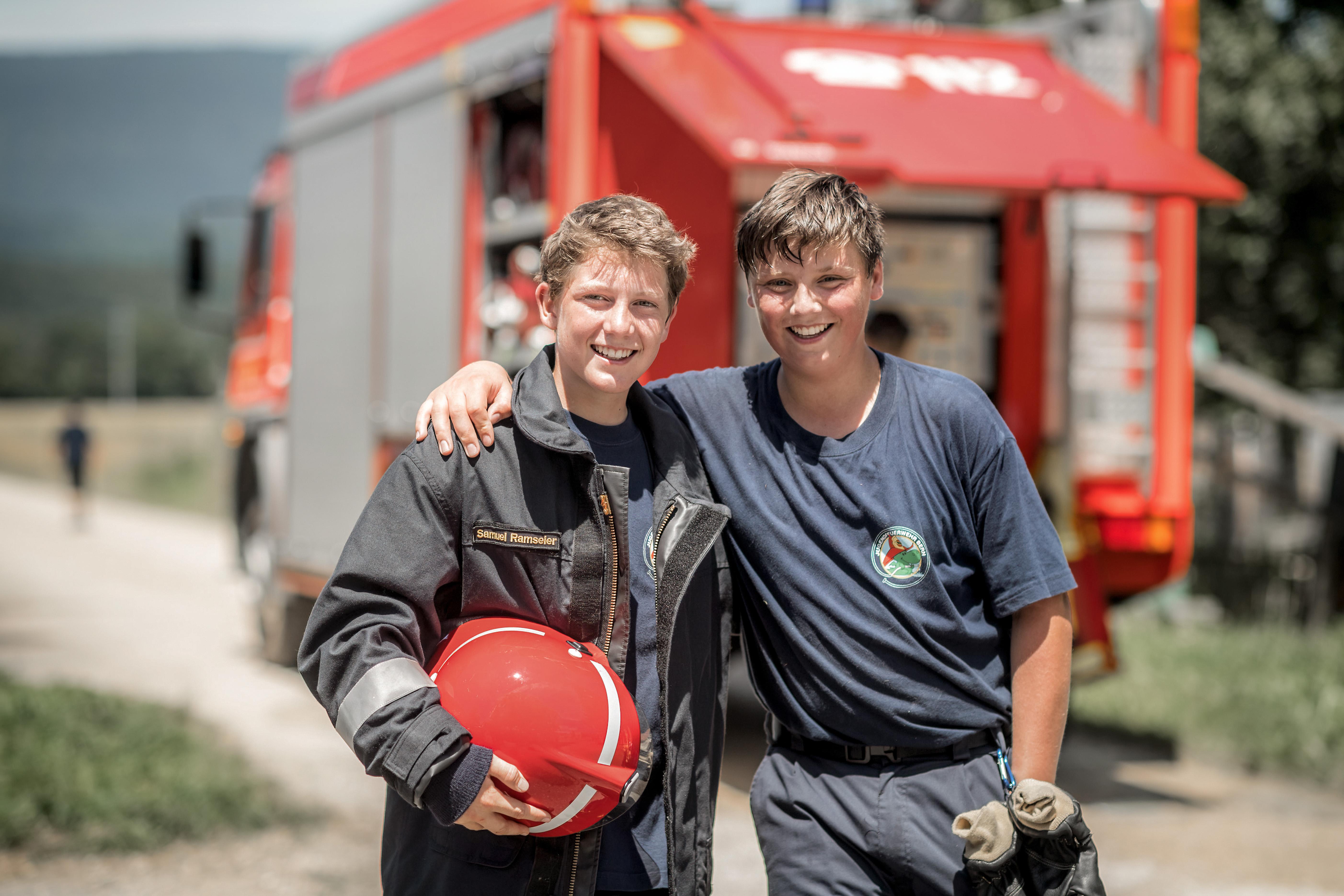  2 junge Feuerwehrmänner mit Held vor einem Feuerwehrauto